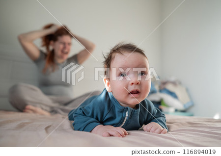 A three-month-old boy lies on his stomach on the bed and his mother sits behind him and cries. Postpartum depression. 116894019