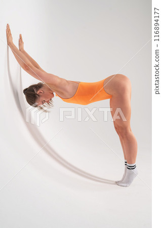 Caucasian woman in orange bodysuit doing downward facing dog on cyclorama. Vertical photo.  116894177