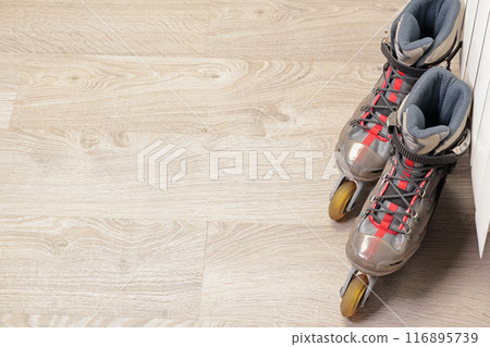 Old roller skates with cloth shoe and plastic base near radiator on wood floor with rubber wheels. Sports and equipment.  116895739