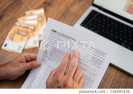 Man company accountant holds tax form examining steps of filling blank correctly sitting at workplace with euro banknotes and laptop with digital table in office 116895836