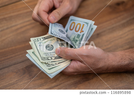 Male company accountant holds dollar banknotes of different denominations carefully counting cash for weekly income report at end of week sitting at wooden table in office Male company accountant holds dollar banknotes of different denominations carefully counting cash for weekly income report at end of week sitting at wooden table in office 116895898