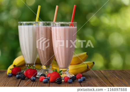 set of milkshakes in glasses with straw and fresh bananas and berries on table outdoors set of milkshakes in glasses with straw and fresh bananas and berries on table outdoors 116897888