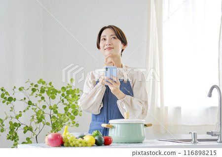 A woman using a smartphone while imagining in the kitchen A woman using a smartphone while imagining in the kitchen 116898215