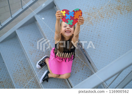 Funny laughing adorable girl shows a heart card made of colorful puzzles as a sign of support for children with autism spectrum disorder 116900562