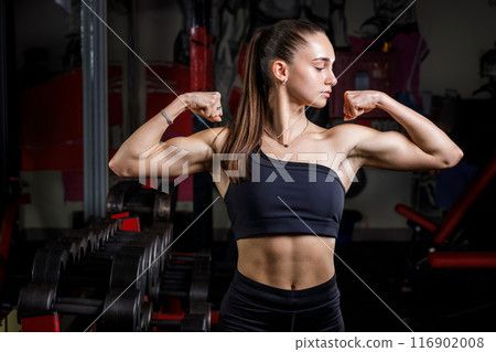 Woman Showing Her Biceps in Gym Near Dumbbell Stand 116902008