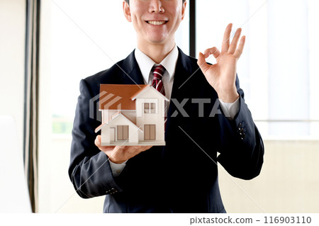 A male business person in a suit holding a house model and saying OK A male business person in a suit holding a house model and saying OK 116903110