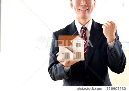 A male business person in a suit holding a house model and making a fist pump 116903380