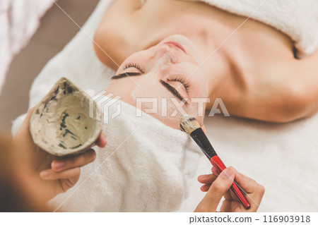 Beautician cosmetologist applies herbal cream mask with spatula, professional procedure in a beauty clinic salon for client. Skincare and cosmetology spa concept. 116903918
