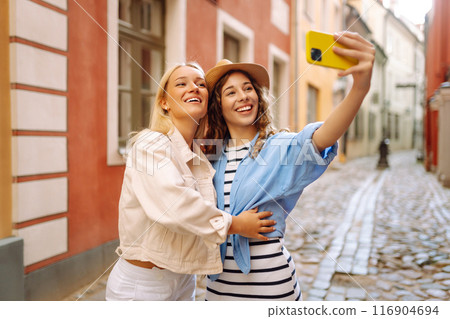 Happy female tourists in casual clothes walk along streets of Europe, take selfies. Active lifestyle 116904694