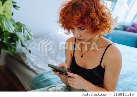 portrait of young woman with curly red hair sitting on bed in bedroom using phone portrait of young woman with curly red hair sitting on bed in bedroom using phone 116911119