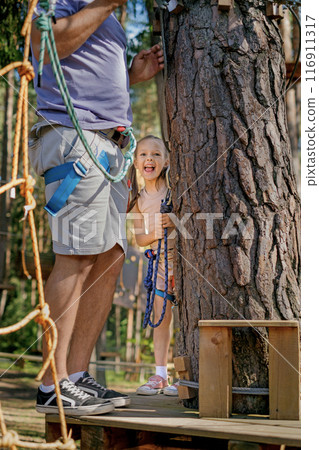 A funny child in full protective gear walks a rope route at a height with his dad A funny child in full protective gear walks a rope route at a height with his dad 116911317