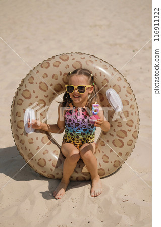 A laughing little girl is sitting in swimming circle on the sand of beach holding birthday cupcake with candle in shape of the number five in hand. Colored swimsuit and lifebuoy with leopard print A laughing little girl is sitting in swimming circle on the sand of beach holding birthday cupcake with candle in shape of the number five in hand. Colored swimsuit and lifebuoy with leopard print 116911322