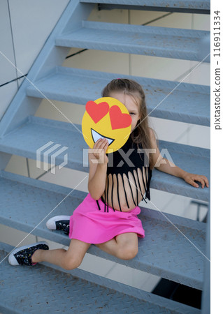 A girl in stylish clothes is sitting on the stairs hiding behind emojis with loving eyes. A way to display emotions through emoticons 116911334