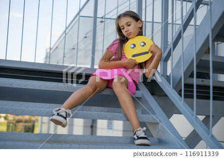 A sad girl holds a Unhappy yellow cardboard Emoticon in her hands while sitting on landing 116911339