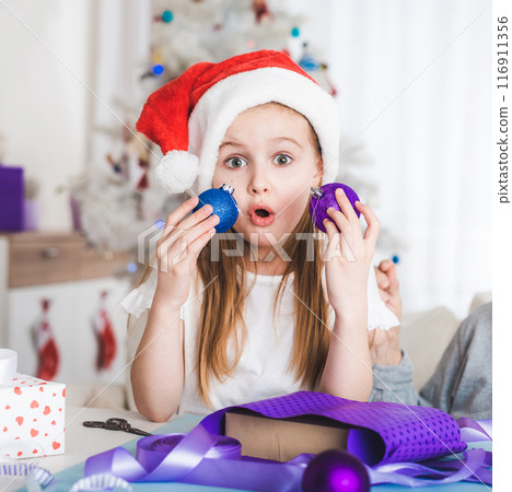 Surprised little girl in santa hat holding blue and purple balls at Christmas Surprised little girl in santa hat holding blue and purple balls at Christmas 116911356