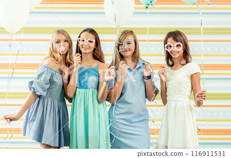 Pretty smiling teenage girls in dresses standing together with ballons at birthday party on the striped colorful background Pretty smiling teenage girls in dresses standing together with ballons at birthday party on the striped colorful background 116911531