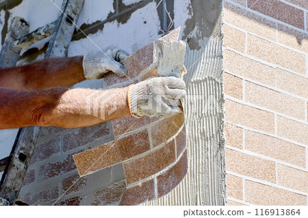 External cladding of facade house with thin faux brick panels, construction worker uses tile adhesive. 116913864