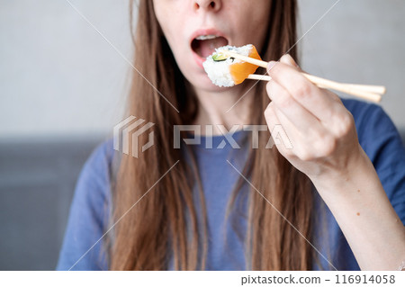 A young woman with long hair holds rolls on sticks against the background of an open mouth, close-up 116914058