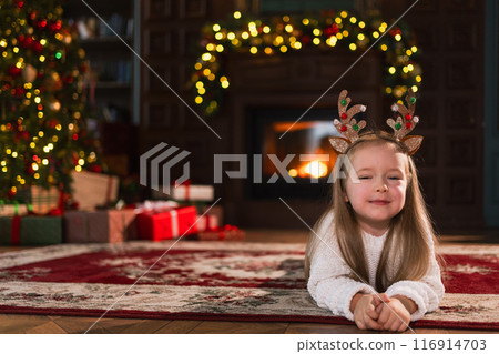 Merry Christmas. Little girl smiling near Christmas tree in classical dark interior. Young happy kid wearing deer horns in living room with fireplace Christmas tree gift boxes. Christmas eve at home 116914703