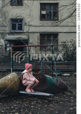 Sad little girl on a pink shabby swing in a bad neighborhood. Sad little girl on a pink shabby swing in a bad neighborhood. 116915010