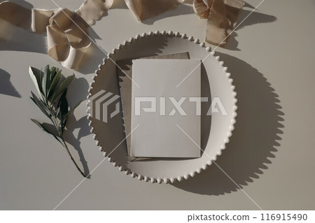 Summer wedding stationery mock-up scene. Blank greeting card, invitation template, white ceramic plate in sunlight. Olive tree branch, silk ribbon on beige table background. Mediterranean flat lay 116915490