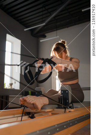 Woman training pilates on the reformer bed. Reformer pilates studio machine for fitness workouts in gym. Fit, healthy and strong authentical body. Fitness concept 116917410