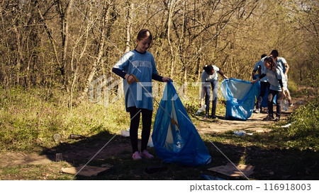 Small girl collecting rubbish around a forest with a claw tool and garbage bags, cleaning up the environment by recycling plastic waste and trash. Child learning to protect ecosystem. Camera B. 116918003