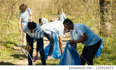 Environmental activists collecting rubbish and plastic waste in garbage bag, using littler cleanup tools to pick up trash from the forest. Women doing voluntary work to protect ecosystem. Camera A. 116918033