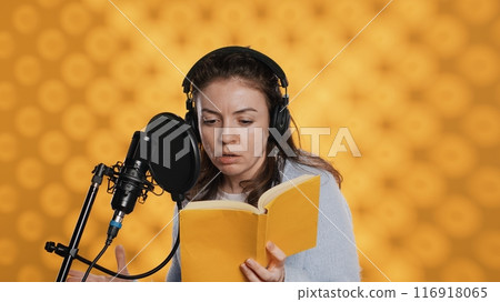 Woman does voiceover reading of book to produce audiobook using dramatic acting. Narrator uses storytelling skills and expressive sounds while producing recording of novel, studio background, camera A 116918065