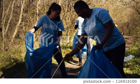 Diverse group of activists gathering to clean up a forest area, using litter cleanup tools like garbage bags and a long claw to grab the trash. People picking rubbish and recycling. Camera B. 116918100