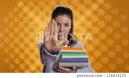 Portrait of stern woman holding stack of books doing stop sign gesturing, studio background. Student with pile of textbooks in arms used for academic learning doing halt hand gesture, camera B 116918135