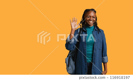 Portrait of chipper girl holding backpack, waving hand, gesturing, isolated over yellow studio background. Jolly african american student with school rucksack doing greeting hand gesture, camera B 116918197