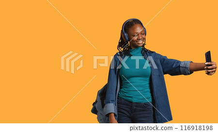 Jolly teenager using cellphone to take selfies doing victory hand sign. Smiling BIPOC girl taking pictures using mobile phone, doing victory gesturing, listening music, studio background, camera B Jolly teenager using cellphone to take selfies doing victory hand sign. Smiling BIPOC girl taking pictures using mobile phone, doing victory gesturing, listening music, studio background, camera B 116918198