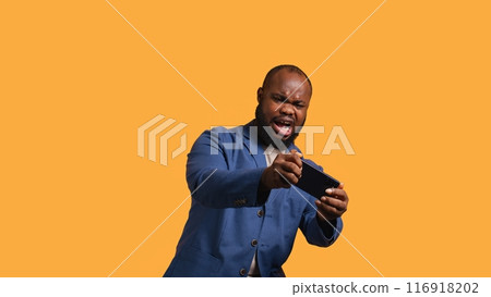 BIPOC person playing videogames on smartphone, having negative burst of emotions after defeat. Frustrated gamer losing game on phone, isolated over studio background, camera B 116918202