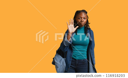 Assertive african american woman doing stop hand gesture and talking. Annoyed girl doing firm halt sign gesturing, wishing to end concept, isolated over studio background, camera B Assertive african american woman doing stop hand gesture and talking. Annoyed girl doing firm halt sign gesturing, wishing to end concept, isolated over studio background, camera B 116918203