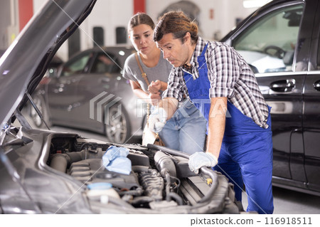 Woman talking to auto mechanic in uniform about repairing his car engine in auto repair shop 116918511