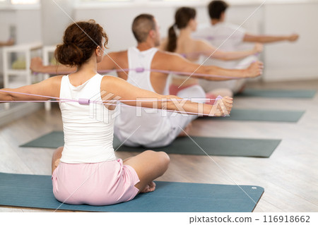 Back view of slim flexible athletic young woman sitting on sports mat practicing pilates during group exercises with elastic band in sports club 116918662