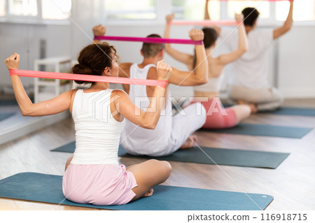Rear view of athletic slim young woman sitting on gymnastics mat performing exercises with elastic band for fitness in gym during group exercise 116918715