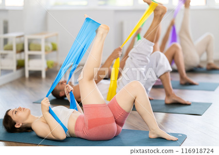 Happy girl and group of women and men lying on mats and doing stretch exercises with resistance bands in fitness room 116918724