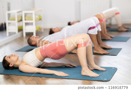 Concentrated girl and group of sporty people lying on mats and performing set of pilates exercises with resistance bands in fitness studio 116918779