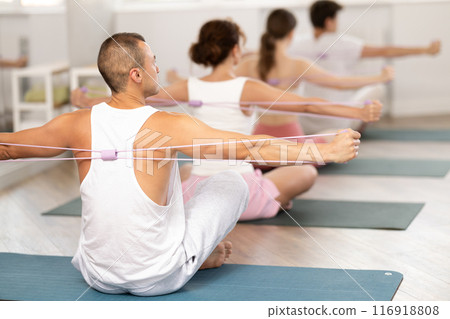 Young guy doing shoulder workout with resistance band sitting cross-legged 116918808