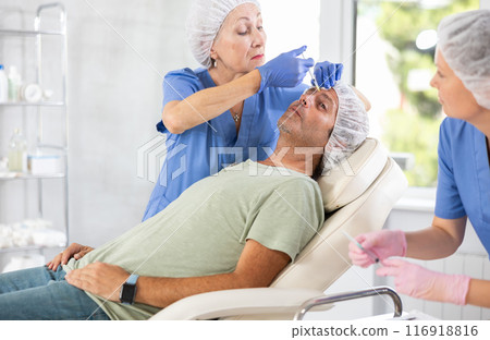 Senior female cosmetologist performing face contouring injections to man 116918816