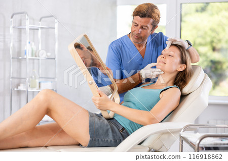 Woman looking at mirror while cosmetologist showing results of facial procedure 116918862