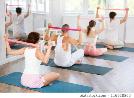 Young men and women doing shoulder workout with resistance band sitting cross-legged 116918863