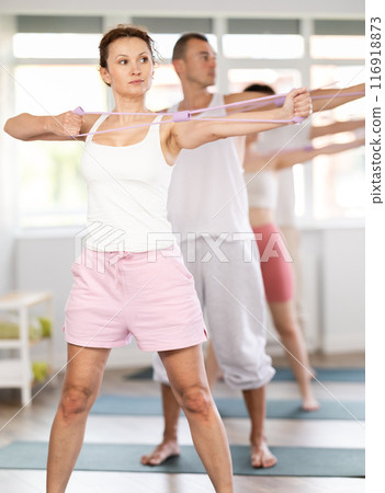 Portrait of young energetic sporty woman standing and perform exercise with resistance bands in pilates studio 116918873