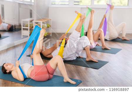 Happy girl and group of women and men lying on mats and doing stretch exercises with resistance bands in fitness room 116918897
