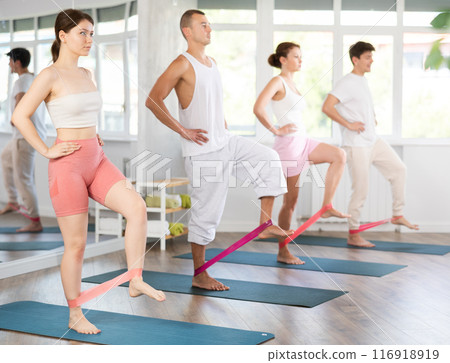 Young men and women practicing hip workout with band standing on mat 116918919