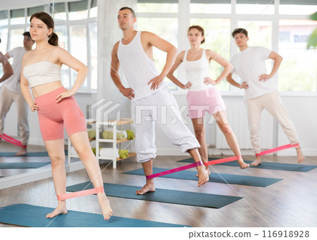 Young men and women practicing hip workout with band standing on mat 116918928