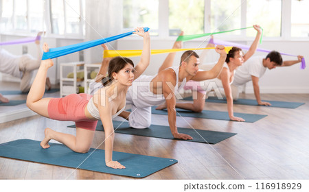 Happy girl on hands and knees doing stretch exercises for legs with resistance band on mats with group of sporty people in gym Happy girl on hands and knees doing stretch exercises for legs with resistance band on mats with group of sporty people in gym 116918929