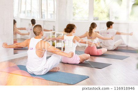 Back view of energetic active young man sitting on gymnastics mat exercising with pilates elastic resistance band with group of athletic people in fitness studio 116918946
